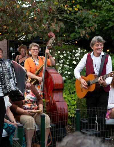 Drie muzikanten in rode vesten spelen accordeon, contrabas en gitaar tijdens een tuinconcert op Piet's 70 party. Foto: AbDonkerFotografie.